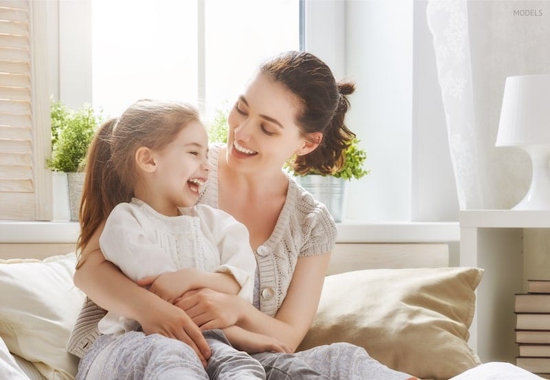 Young mother wrapping her arms around her young daughter, sitting on bed