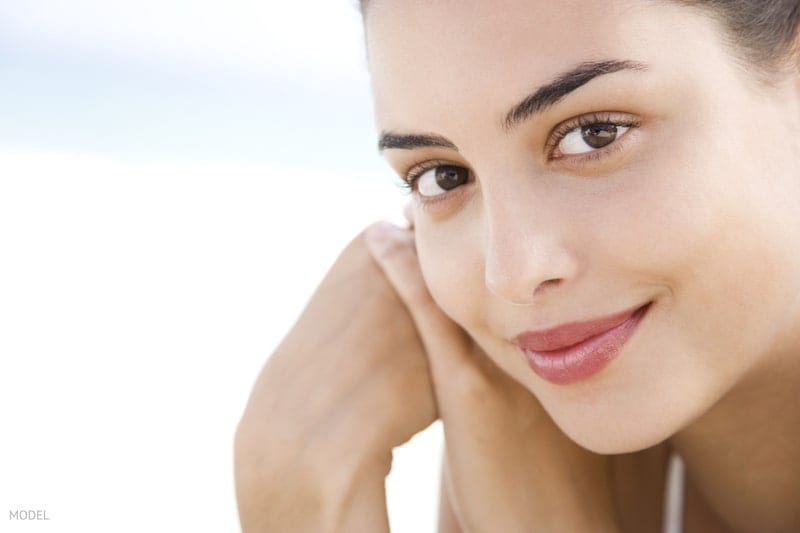 Close-up of beautiful young woman's face looking at camera with her hands clasped by her cheek. 