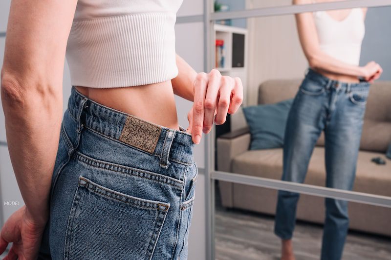 Woman admiring her contoured body in the mirror as she wears jeans that are too big for her