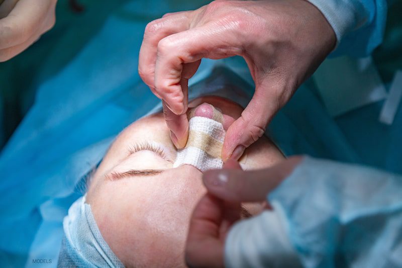 Woman on the operating table while a cosmetic surgeon puts a bandage on her nose.